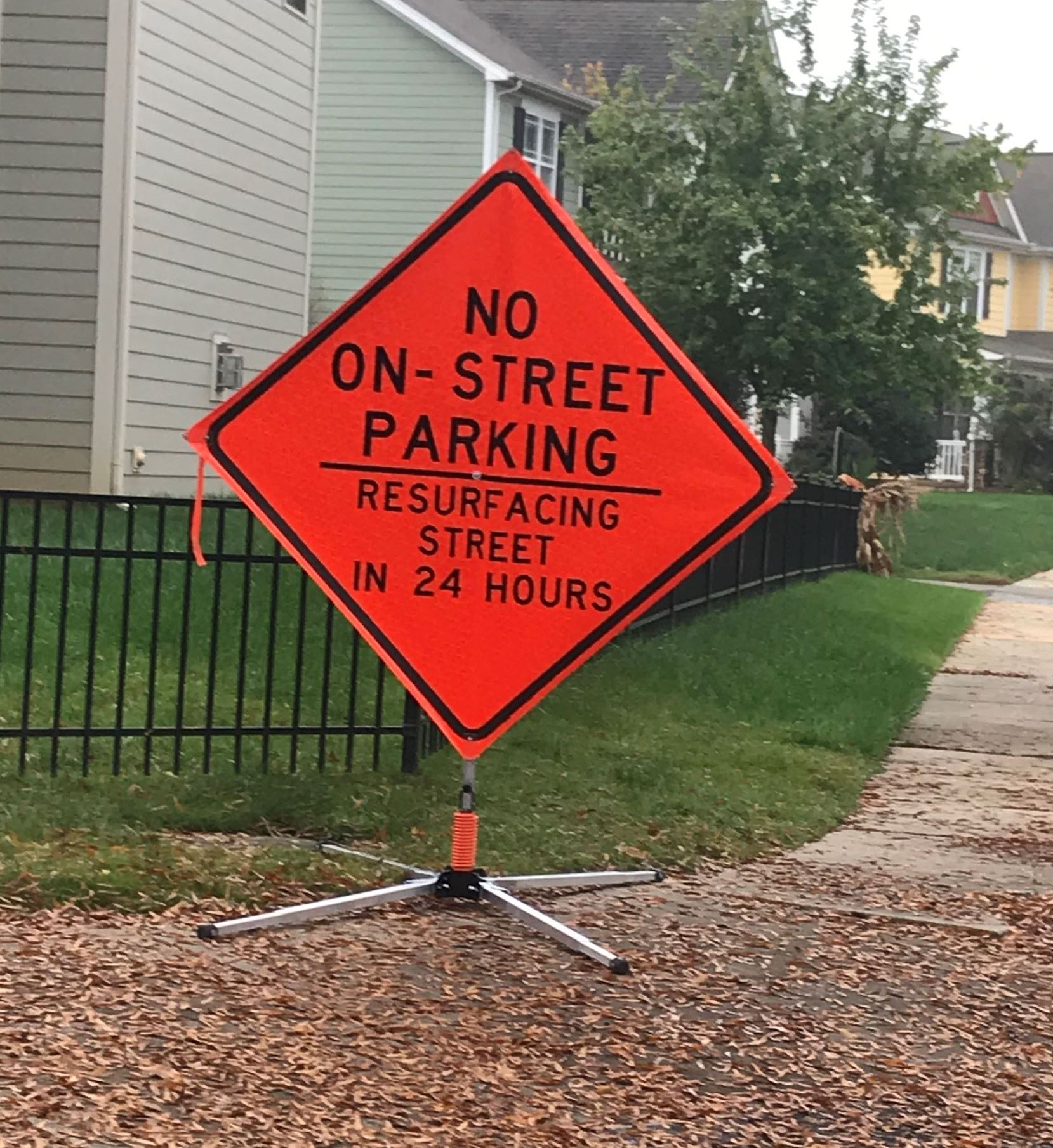 An orange No On-Street Parking sign sits on a sidewalk next to the back of a house.